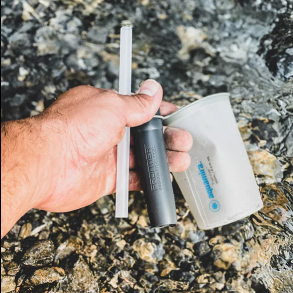 Hand holding an Aquamira Frontier Straw filter and storage pouch beside a rocky stream during an outdoor adventure.