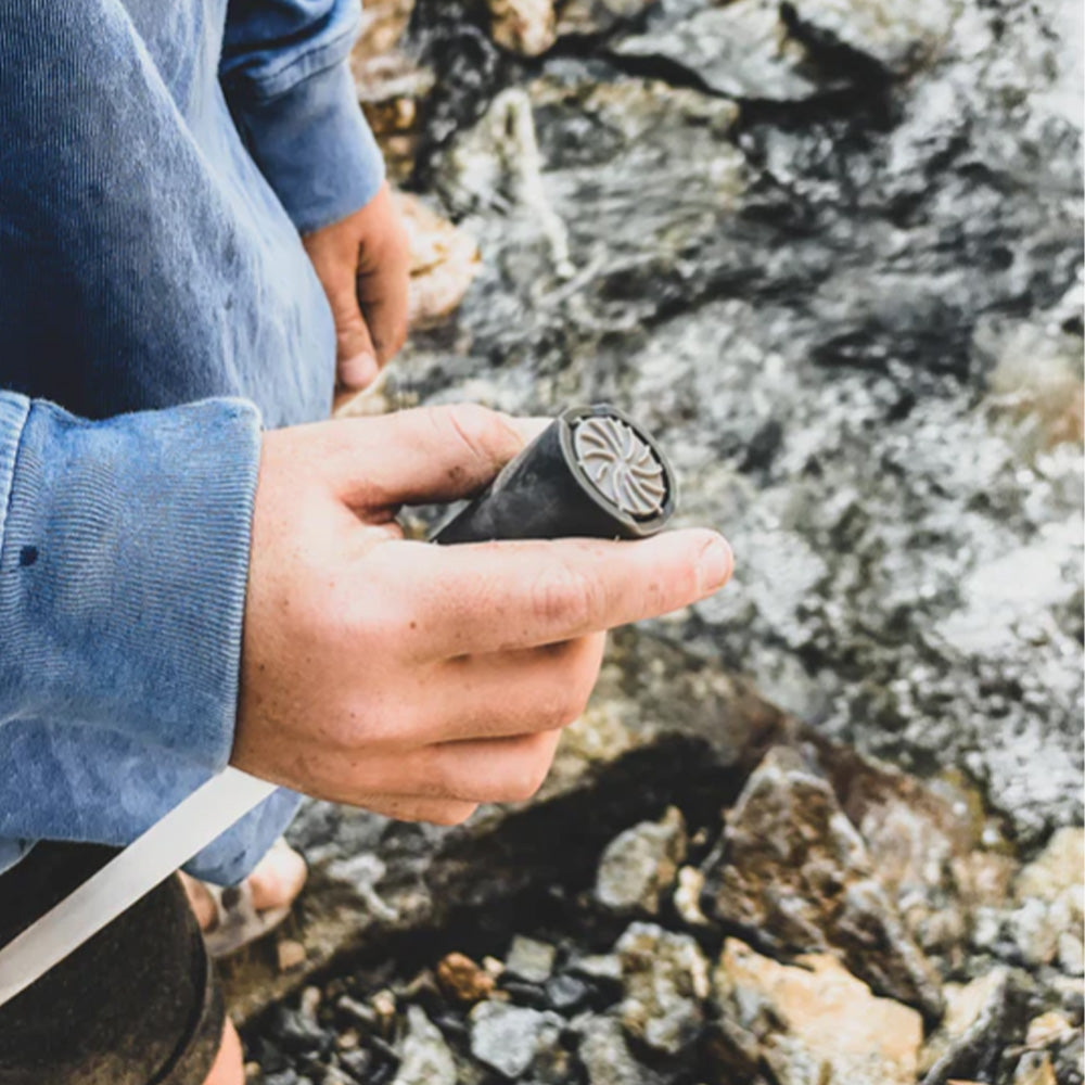 Person handling an Aquamira Frontier Straw portable water filter while seated outdoors near a rocky stream.