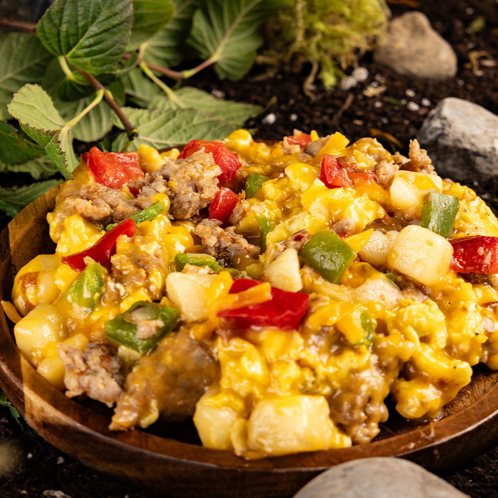 A wood bowl with a helping of scrambled eggs, vegetables, meat, and cheese. Dirt, rocks, and leaves in the background.