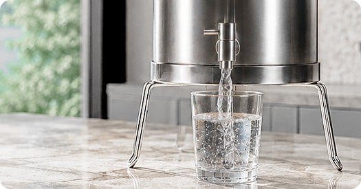 A stainless steel water dispenser with a metal spout pouring clear water into a clear glass tumbler, set on a marble countertop in a kitchen environment.