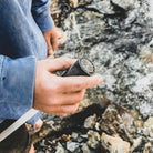 Person handling an Aquamira Frontier Straw portable water filter while seated outdoors near a rocky stream.