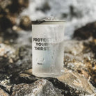 Aquamira Frontier Straw storage pouch standing upright on rocks beside flowing water in an outdoor setting.