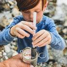 Person drinking water through an Aquamira Frontier Straw portable filter while outdoors near a rocky stream.