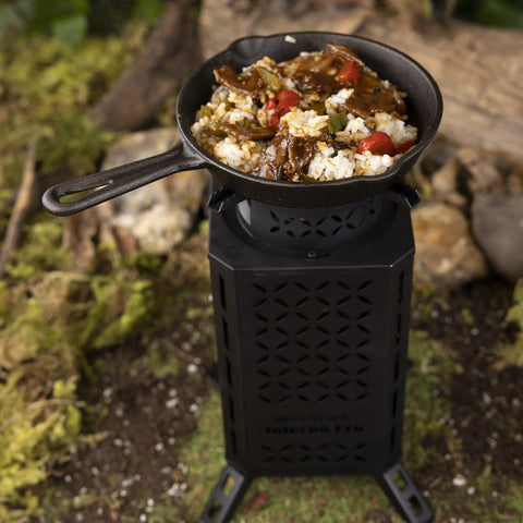 Image of A cast iron pan with rice, meat, and vegetables cooking on top of the Inferno Pro stove, sitting in a forest of dirt and grass.