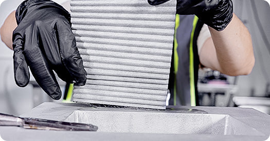 A close-up of a person wearing black gloves handling a gray pleated filter or mesh panel over a white tray in a workshop or industrial setting.