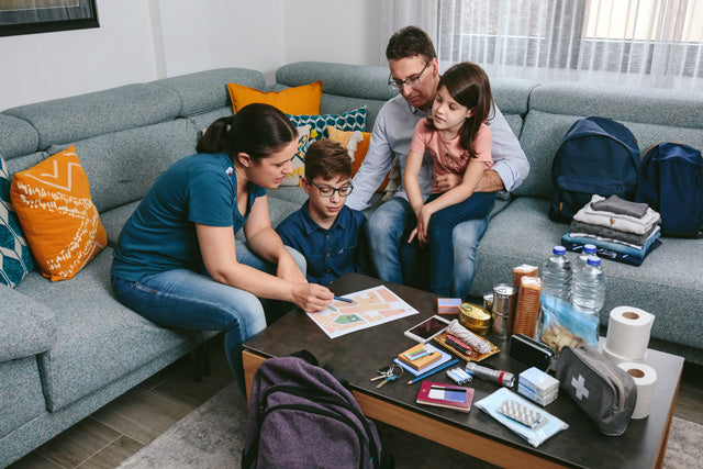 Family of four sitting on a couch in a living room with a table full of items.