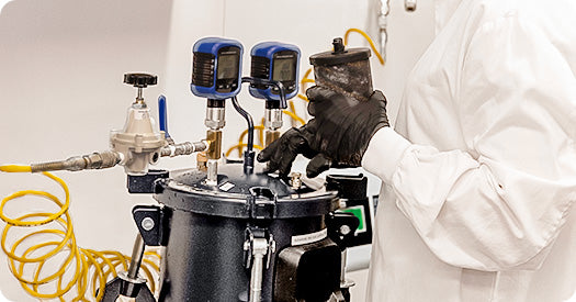 A person in a white lab coat and black gloves adjusts a piece of industrial laboratory equipment with two blue digital gauges, yellow hoses, and metal valves in a clean indoor setting.