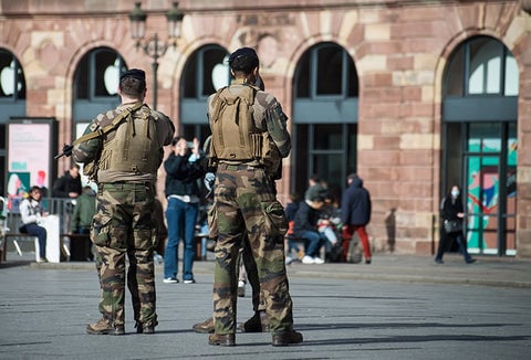 Military men patrolling a street of civilians with weapons in hand.