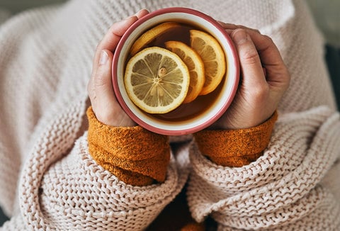 A person wrapped in a blanket holding a hot drink with a lemon in it.