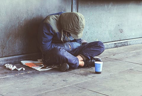 A homeless person in a coat sitting against a concrete wall, with food scraps and garbage around him.