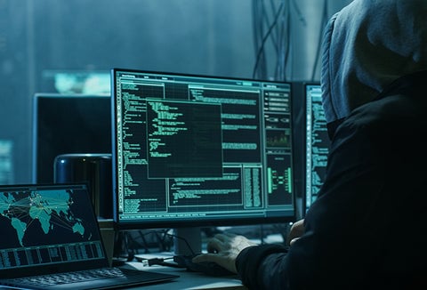A hooded individual in a dark room, hacking into computers.