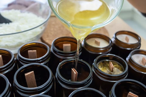 Cooking oil being poured into dark, glass jars with wood wicks to make candles.