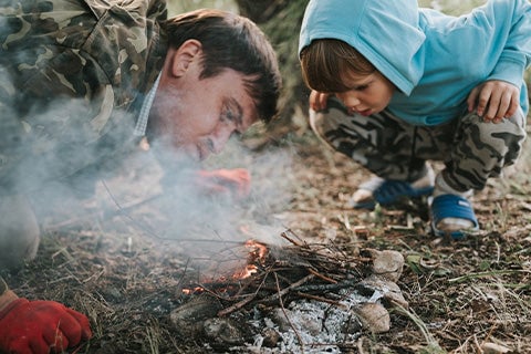 A father and son dressed in outdoor gear, blowing on a fire to help the flames grow.