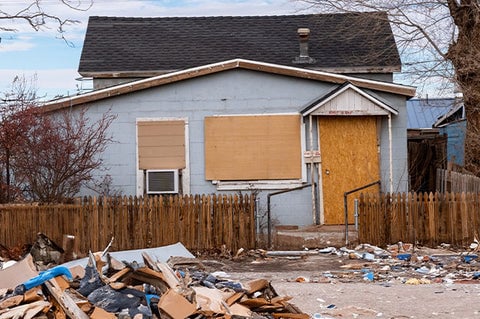 A blue home with boarded-up windows and a pile of trash in the front yard.