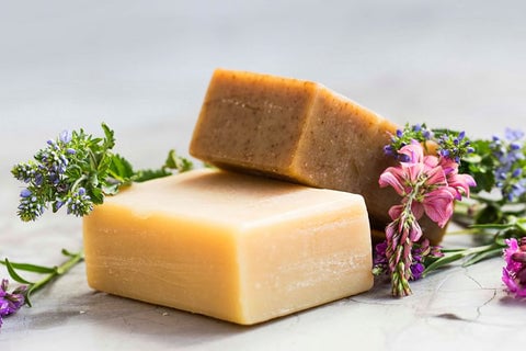 Two beige bars of lye soap made from recycled cooking oil, surrounded by pink and purple flower sprigs.